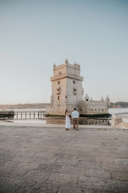 A wide waterfront view at Belém | Lisbon anniversary photoshoot A wide view of the couple walking on the stone paving with Belém Tower and the water behind them during their anniversary photoshoot in Lisbon.