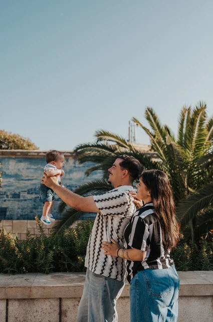 The father lifting the baby gently in front of him with the mother beside them at the tiled viewpoint during their family photoshoot in Lisbon.