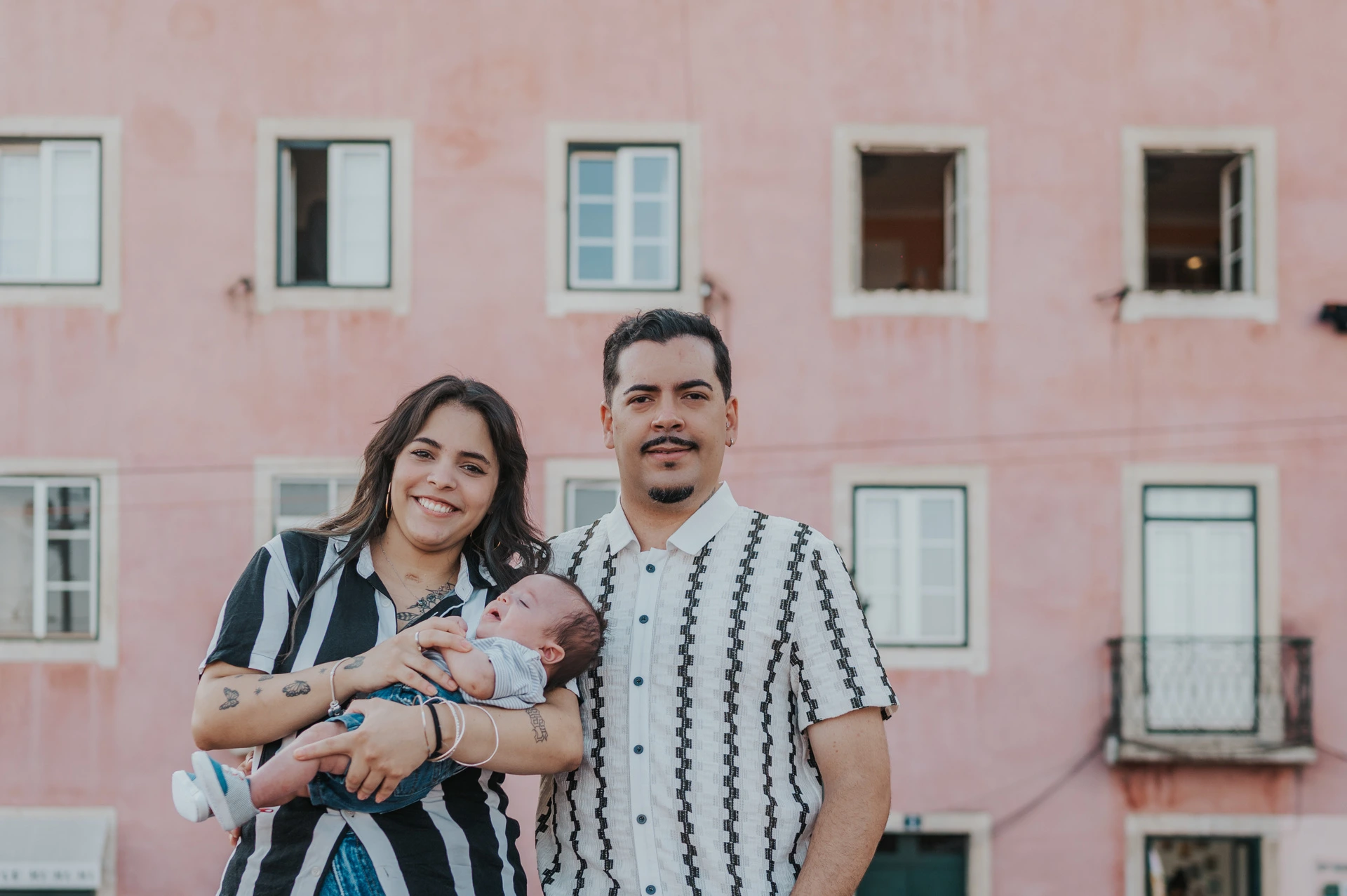 Together against a pink city backdrop | Lisbon family photoshoot The parents holding their baby together with a pink Lisbon building behind them during their family photoshoot in Lisbon.
