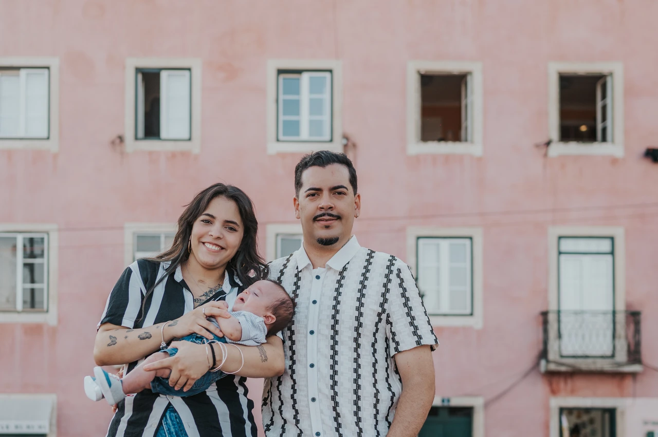 Together against a pink city backdrop | Lisbon family photoshoot The parents holding their baby together with a pink Lisbon building behind them during their family photoshoot in Lisbon.