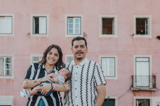 The parents holding their baby together with a pink Lisbon building behind them during their family photoshoot in Lisbon.