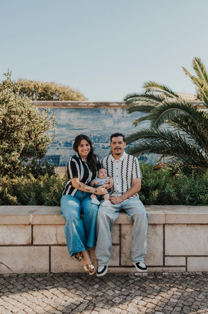 The parents seated together holding their baby beside palm leaves and blue Portuguese tiles during their family photoshoot in Lisbon.