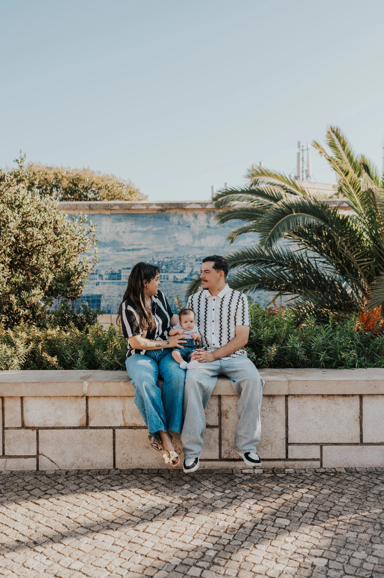 The parents seated together holding their baby beside palm leaves and blue Portuguese tiles during their family photoshoot in Lisbon.