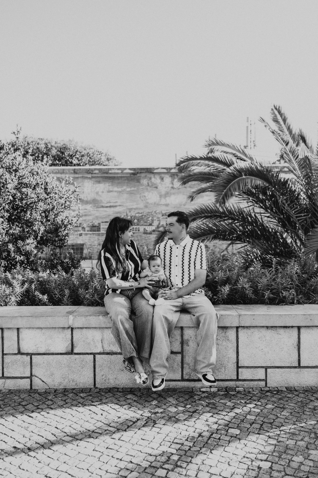Black-and-white photograph: the parents seated together holding their baby beside palm leaves and blue Portuguese tiles during their family photoshoot in Lisbon.