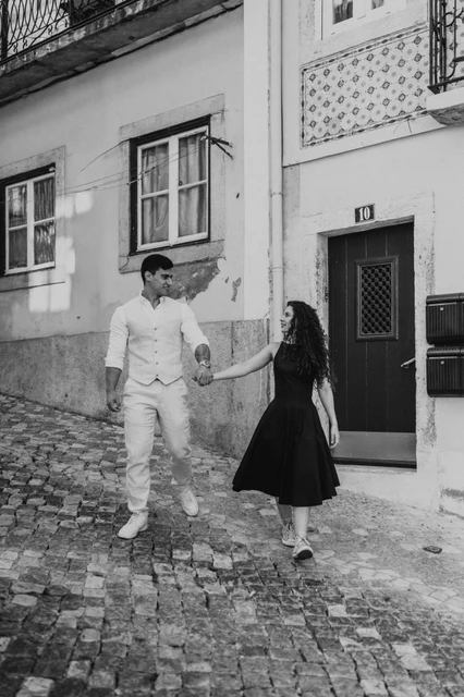 A walk along Bica’s cobbled street in black and white | Lisbon couple photoshoot Black-and-white photograph: the couple walking hand in hand along the cobbled street beside yellow façades during their couple photoshoot in Lisbon.