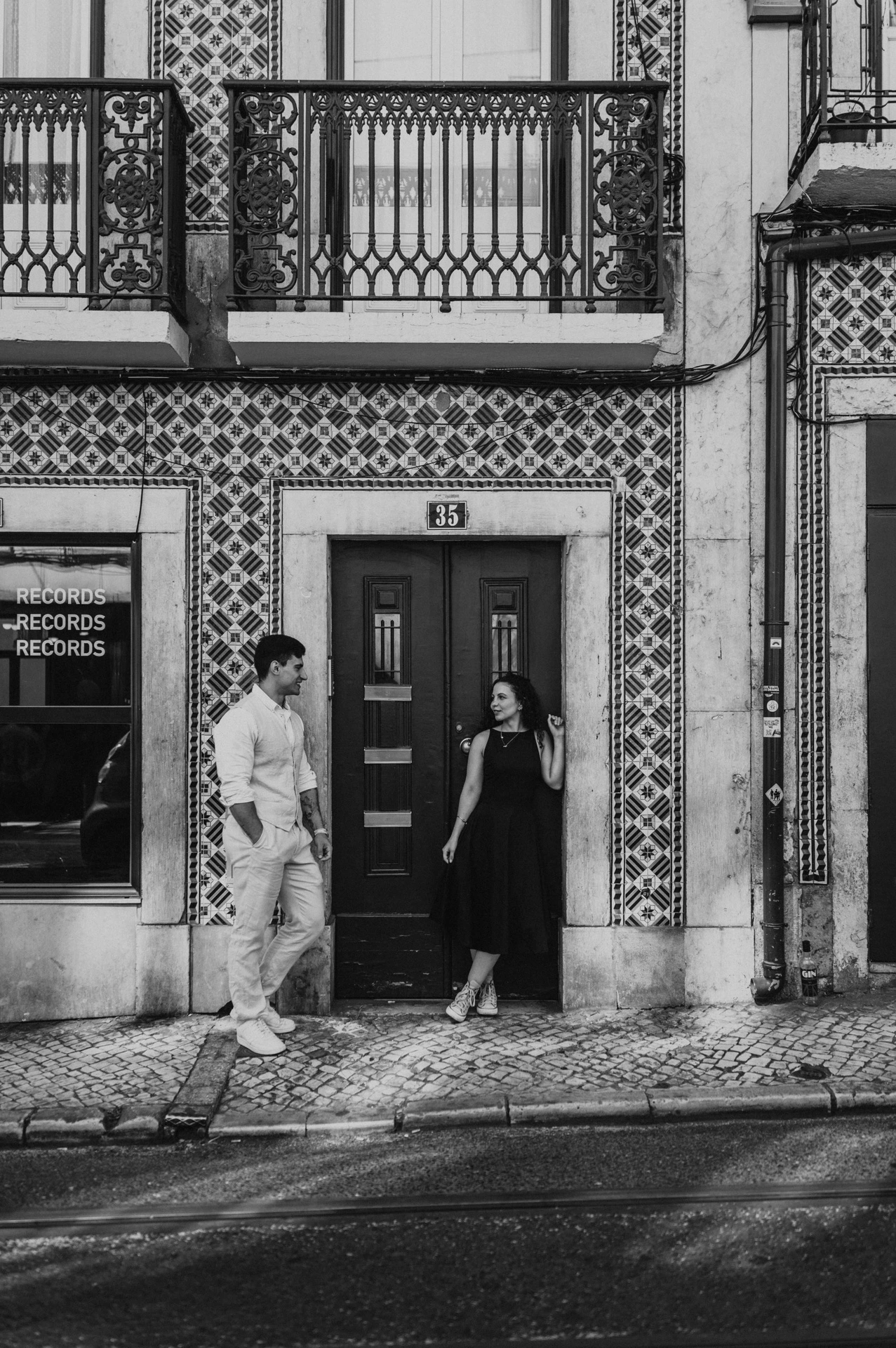 Black-and-white photograph: the couple standing apart beside a green door surrounded by patterned tiles during their couple photoshoot in Lisbon.