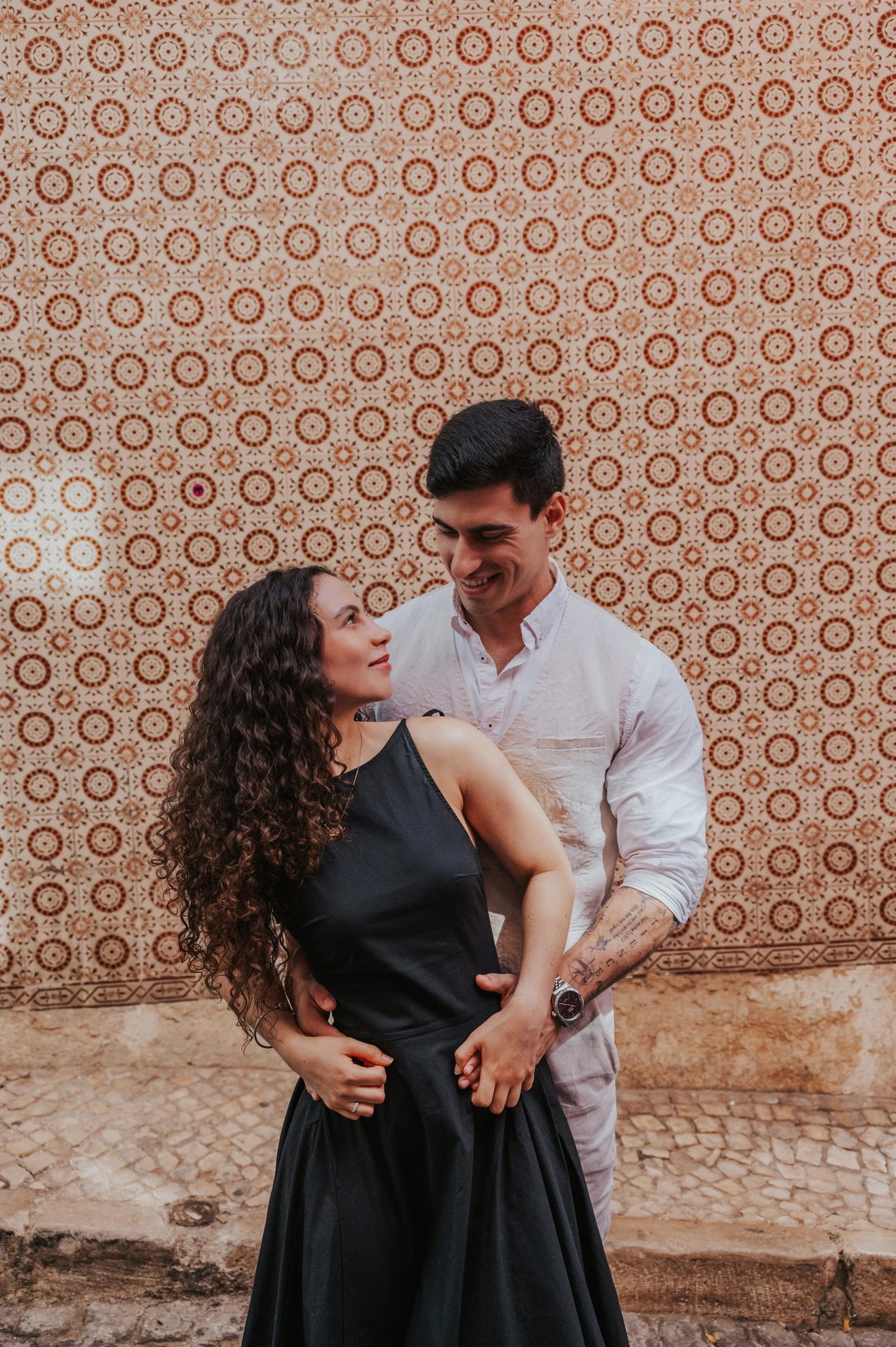 The man embracing his partner from behind against the pink tile wall during their couple photoshoot in Lisbon.