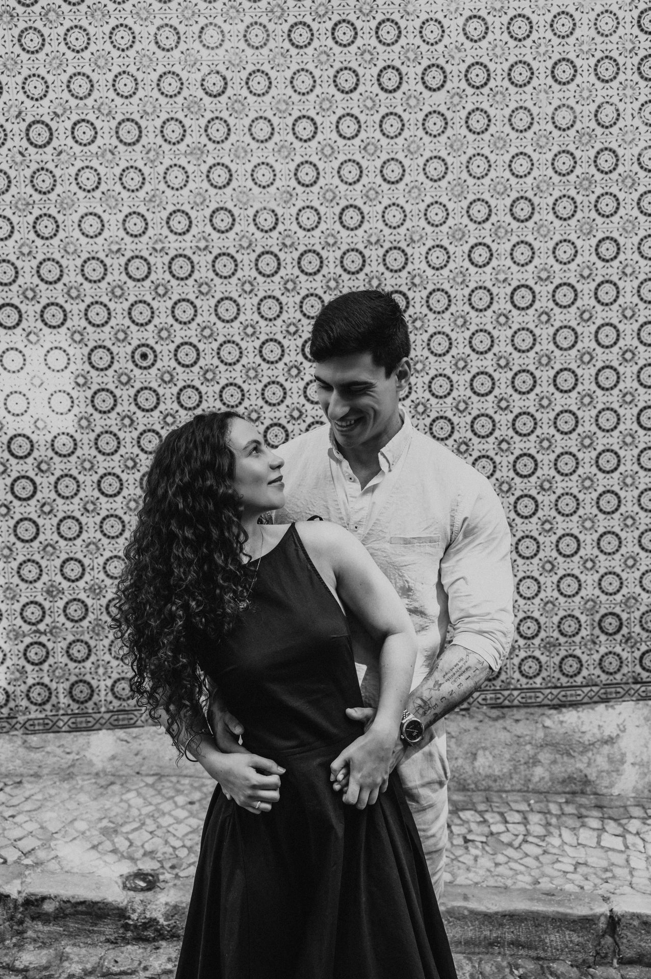 Black-and-white photograph: the man embracing his partner from behind against the pink tile wall during their couple photoshoot in Lisbon.