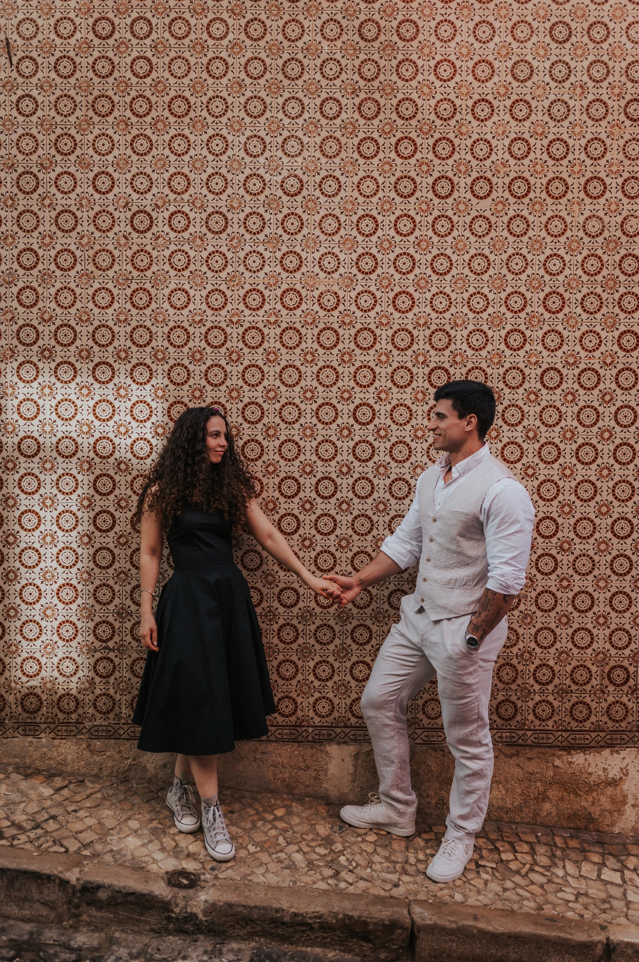 The couple holding hands and looking at one another beside the pink tile wall during their couple photoshoot in Lisbon.