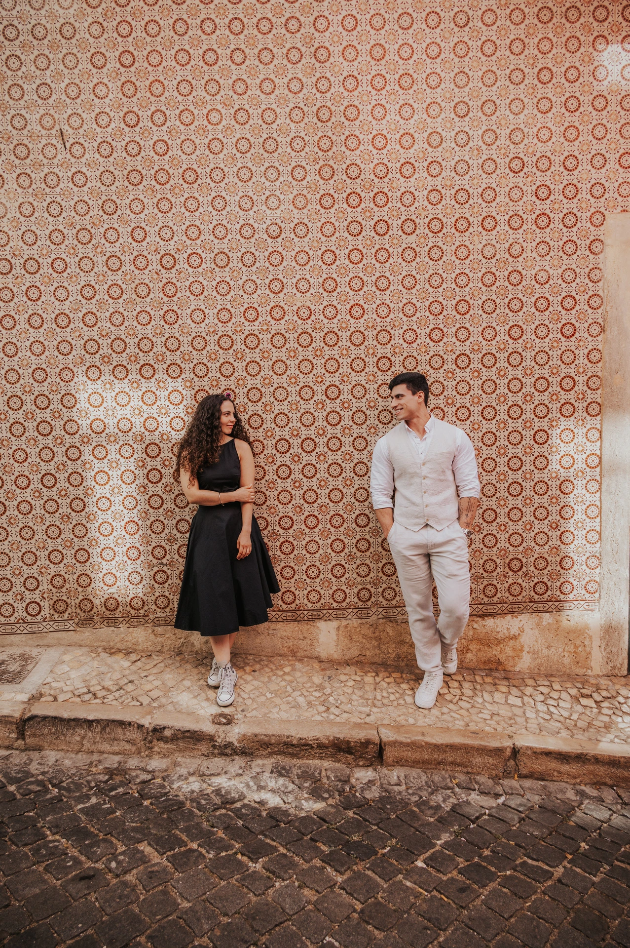 The couple smiling at each other while standing apart against a pink patterned tile wall during their couple photoshoot in Lisbon.