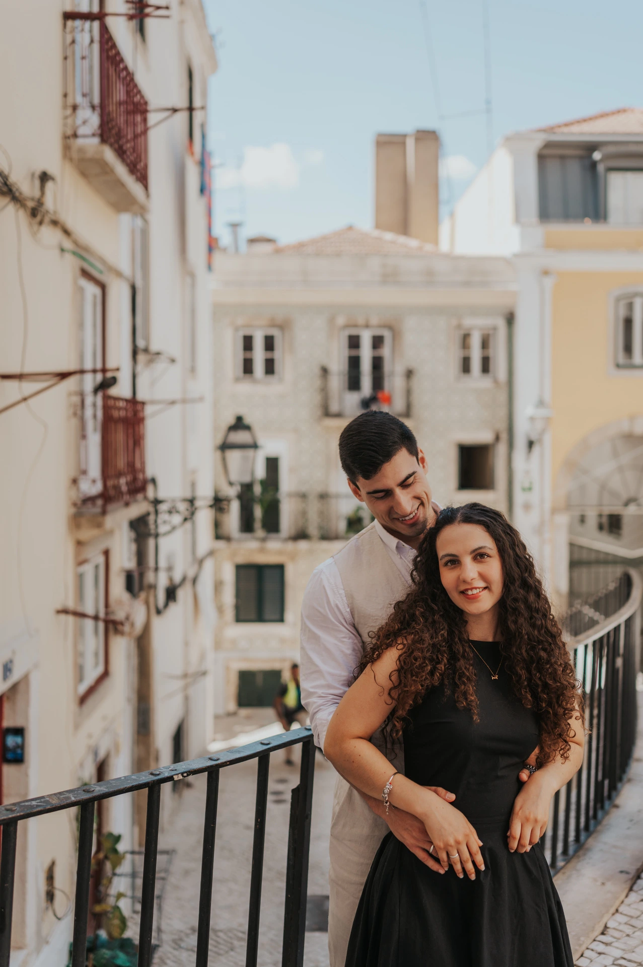 The man embracing his partner from behind beside a railing above the street during their couple photoshoot in Lisbon.