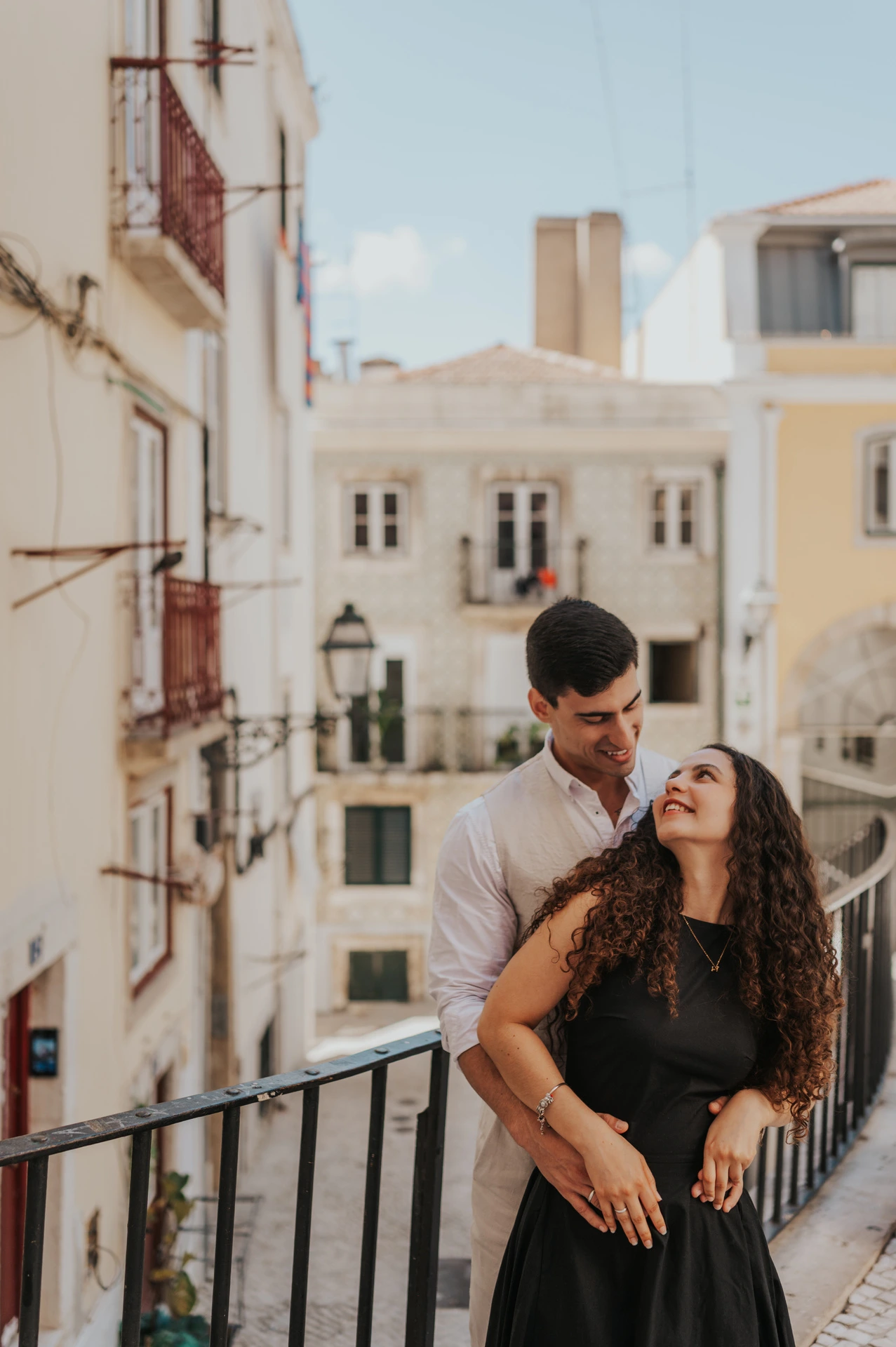 The man embracing his partner from behind beside a railing above the street during their couple photoshoot in Lisbon.