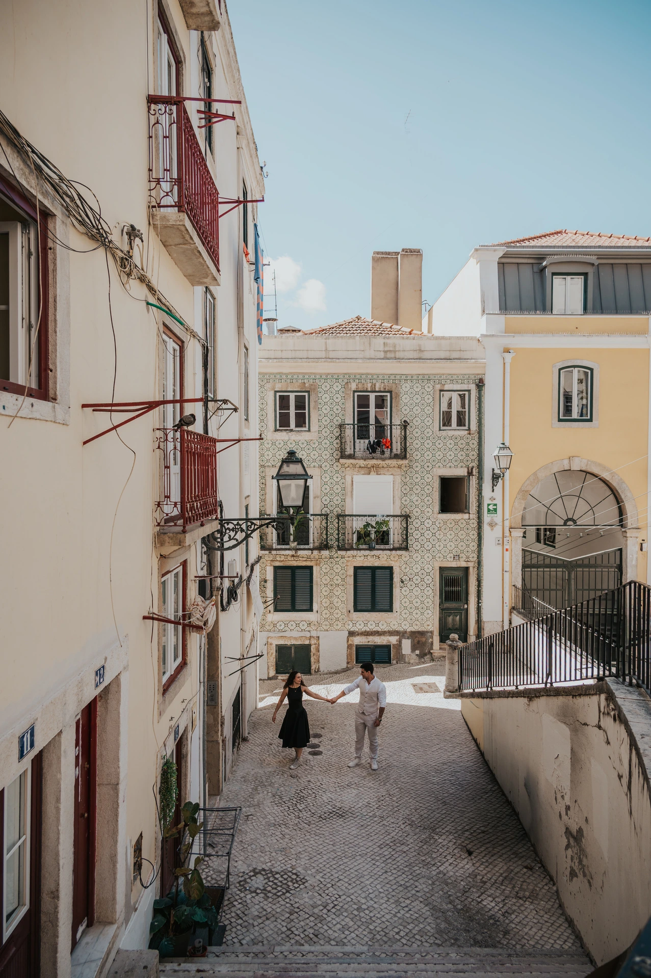 A wide view of the couple beside the railing with the old street and buildings below them during their couple photoshoot in Lisbon.
