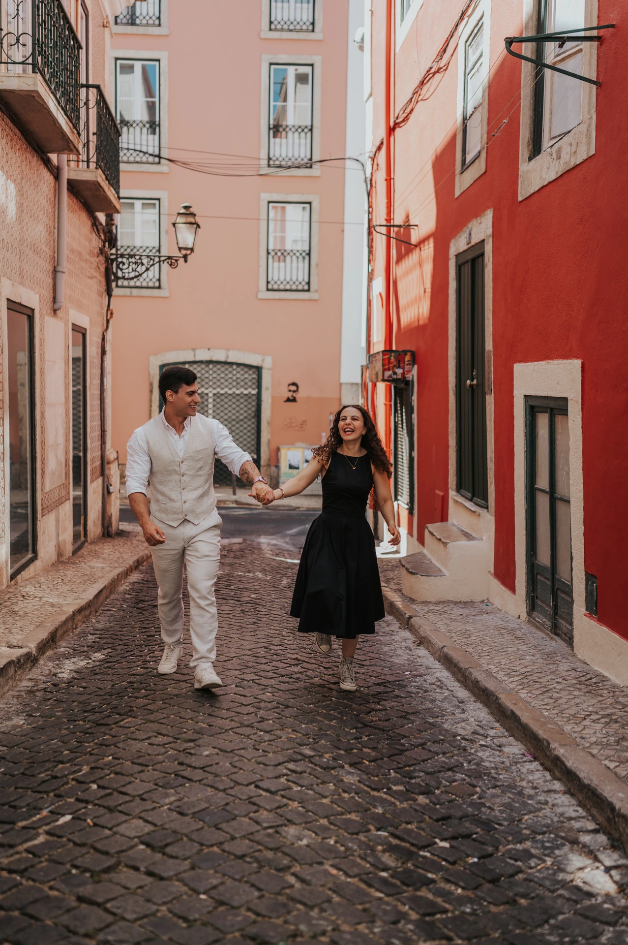The couple smiling and holding hands as they walk together along a narrow street with red façades during their couple photoshoot in Lisbon.