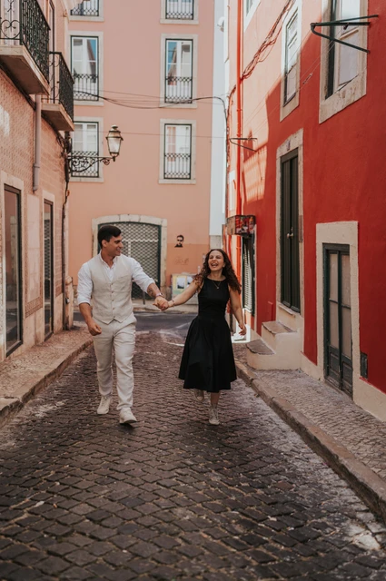 A smiling walk through the colourful street | Lisbon couple photoshoot The couple smiling and holding hands as they walk together along a narrow street with red façades during their couple photoshoot in Lisbon.
