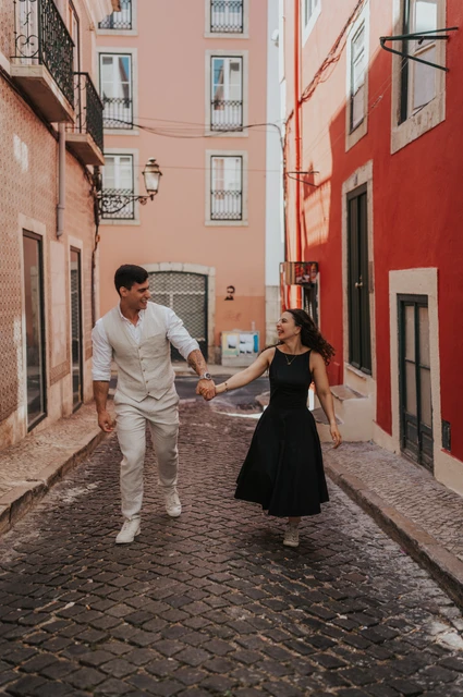 A smiling walk through the colourful street | Lisbon couple photoshoot The couple smiling and holding hands as they walk together along a narrow street with red façades during their couple photoshoot in Lisbon.