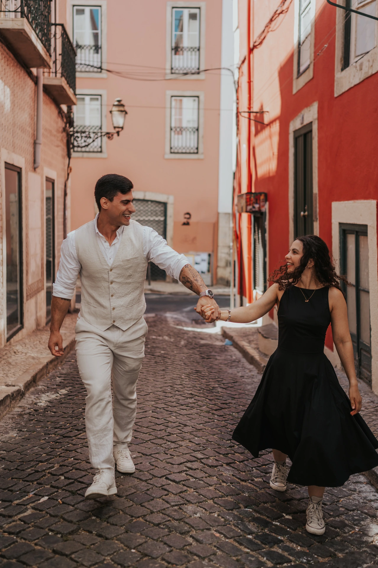 The couple smiling and holding hands as they walk together along a narrow street with red façades during their couple photoshoot in Lisbon.