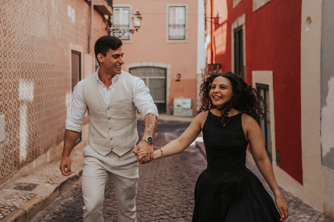 A smiling walk through the colourful street | Lisbon couple photoshoot The couple smiling and holding hands as they walk together along a narrow street with red façades during their couple photoshoot in Lisbon.