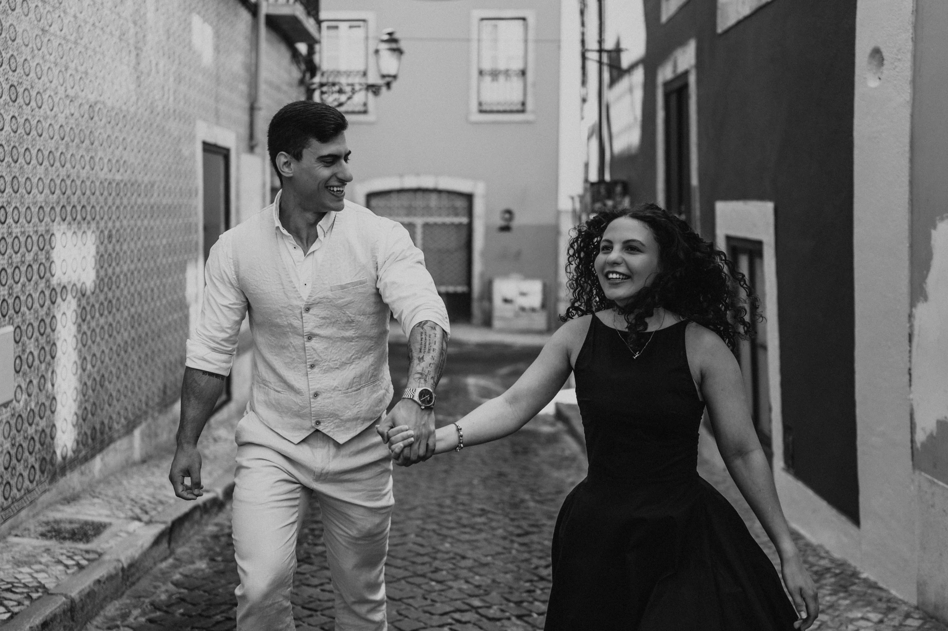 Black-and-white photograph: the couple smiling and holding hands as they walk together along a narrow street with red façades during their couple photoshoot in Lisbon.