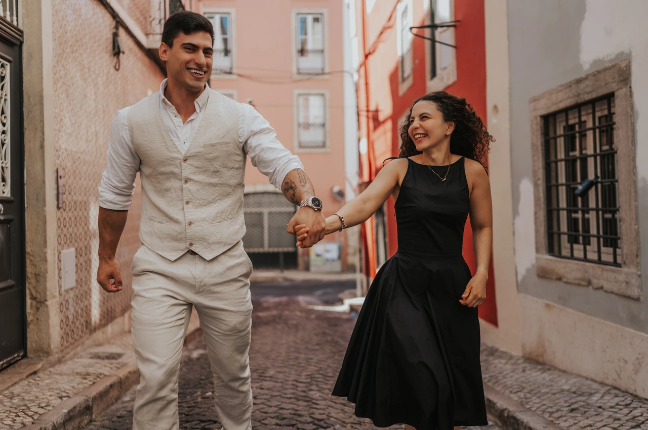 A smiling walk through the colourful street | Lisbon couple photoshoot The couple smiling and holding hands as they walk together along a narrow street with red façades during their couple photoshoot in Lisbon.