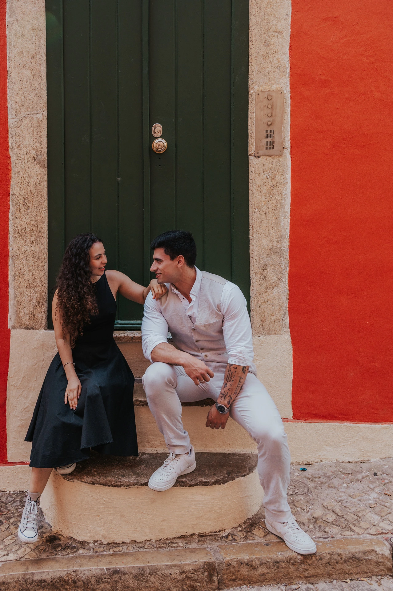 The woman seated on the steps of a green door while her partner stands close beside her against a red wall during their couple photoshoot in Lisbon.