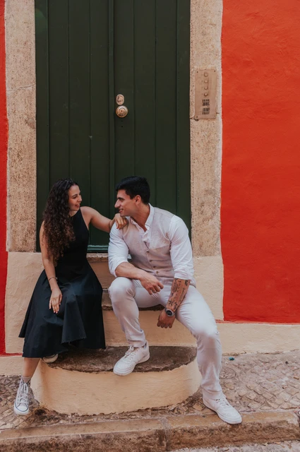 The woman seated on the steps of a green door while her partner stands close beside her against a red wall during their couple photoshoot in Lisbon.