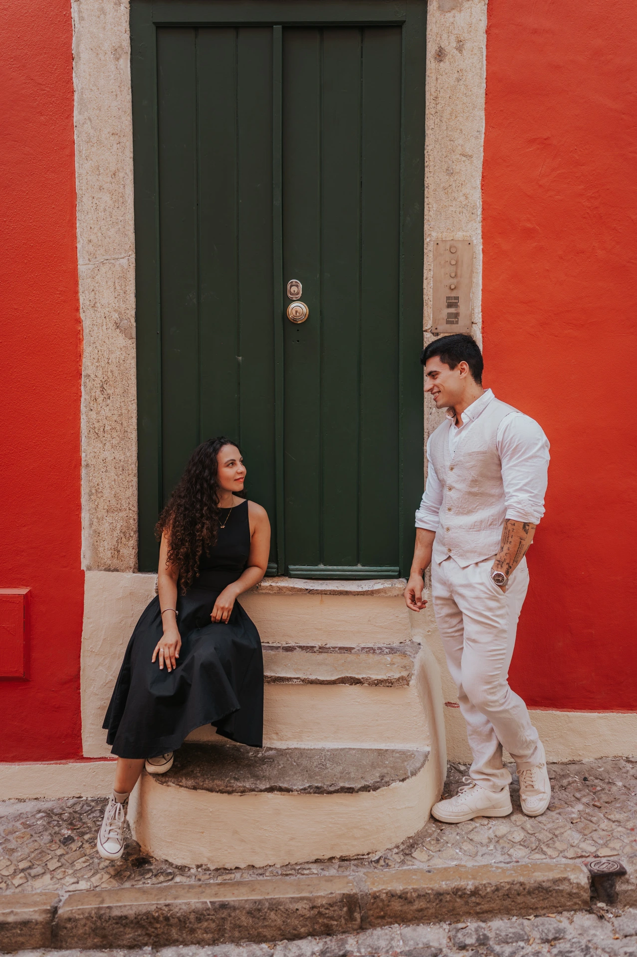 The woman seated on the steps of a green door while her partner stands close beside her against a red wall during their couple photoshoot in Lisbon.