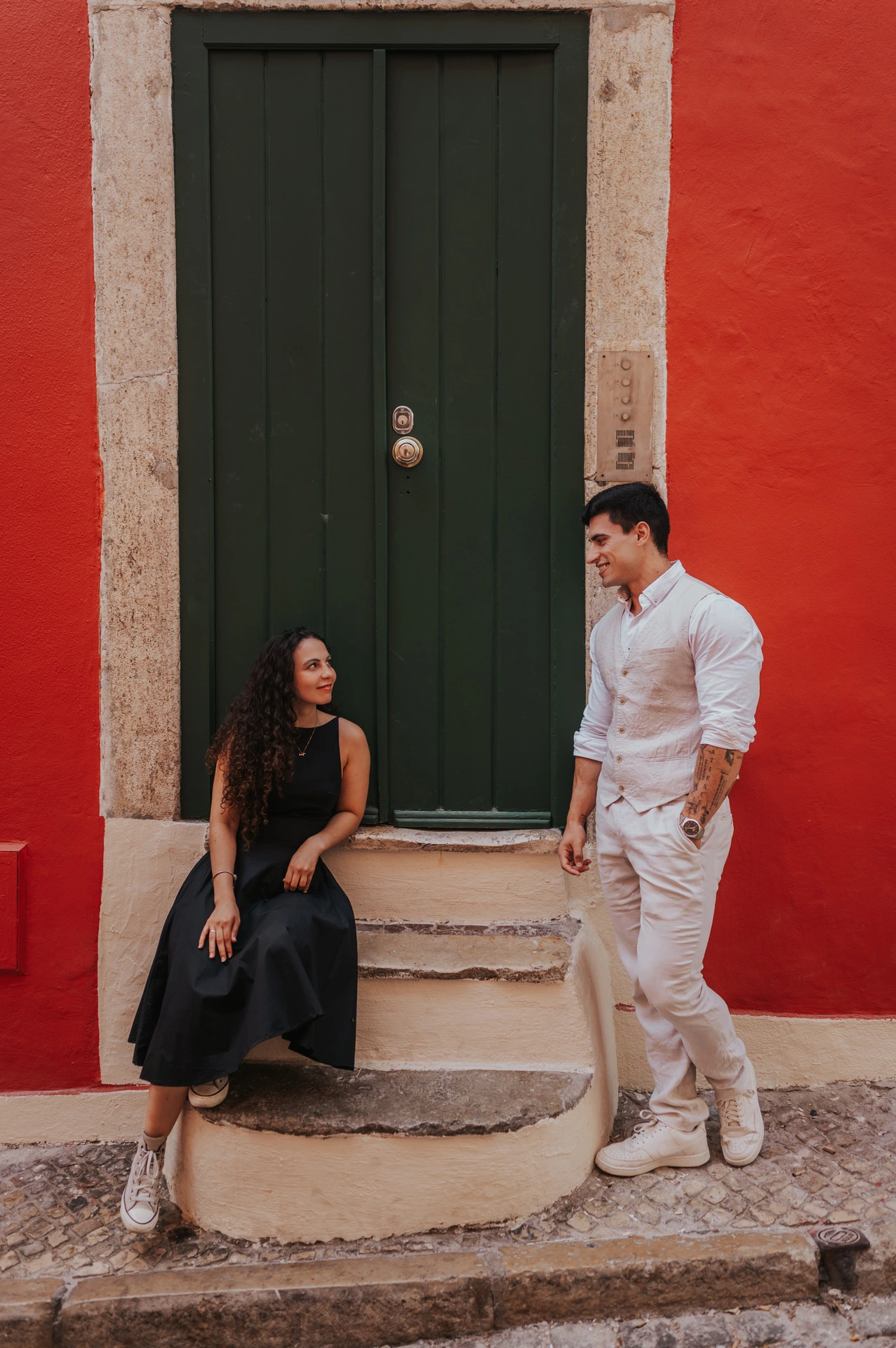 The woman seated on the steps of a green door while her partner stands close beside her against a red wall during their couple photoshoot in Lisbon.