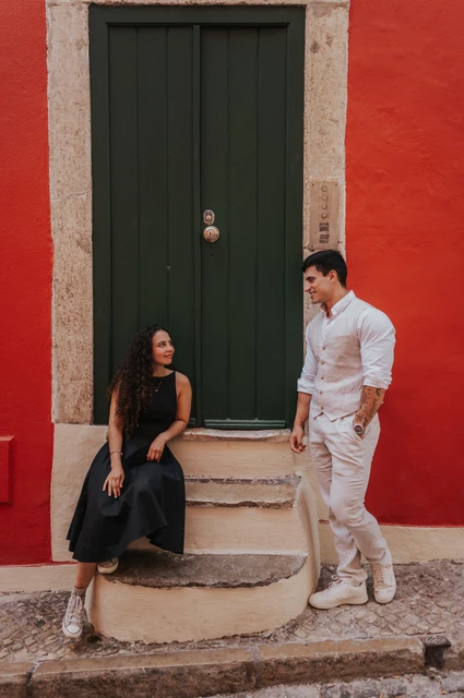 A seated portrait at the red wall | Lisbon couple photoshoot The woman seated on the steps of a green door while her partner stands close beside her against a red wall during their couple photoshoot in Lisbon.