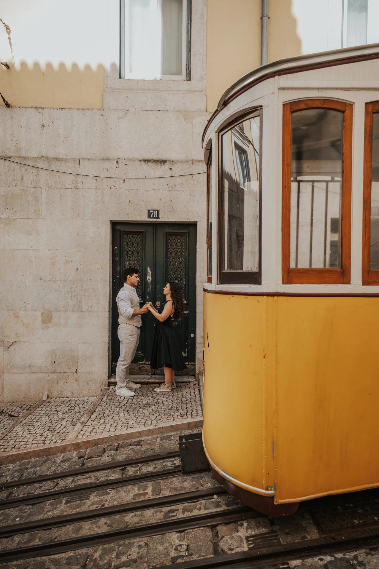 The couple standing close by a doorway with a yellow funicular in the foreground during their couple photoshoot in Lisbon.