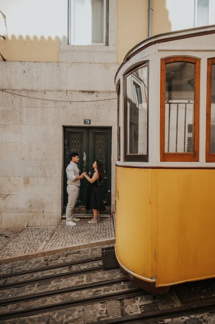 The couple standing close by a doorway with a yellow funicular in the foreground during their couple photoshoot in Lisbon.