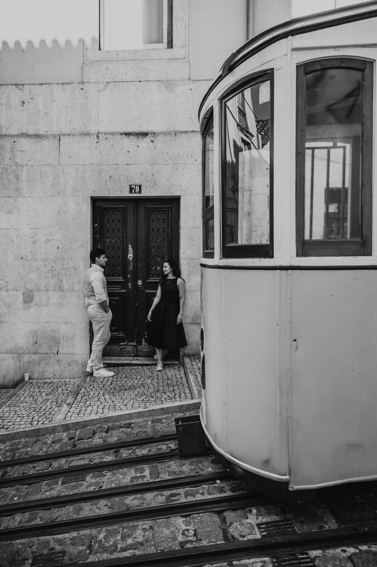 Black-and-white photograph: the couple standing close by a doorway with a yellow funicular in the foreground during their couple photoshoot in Lisbon.