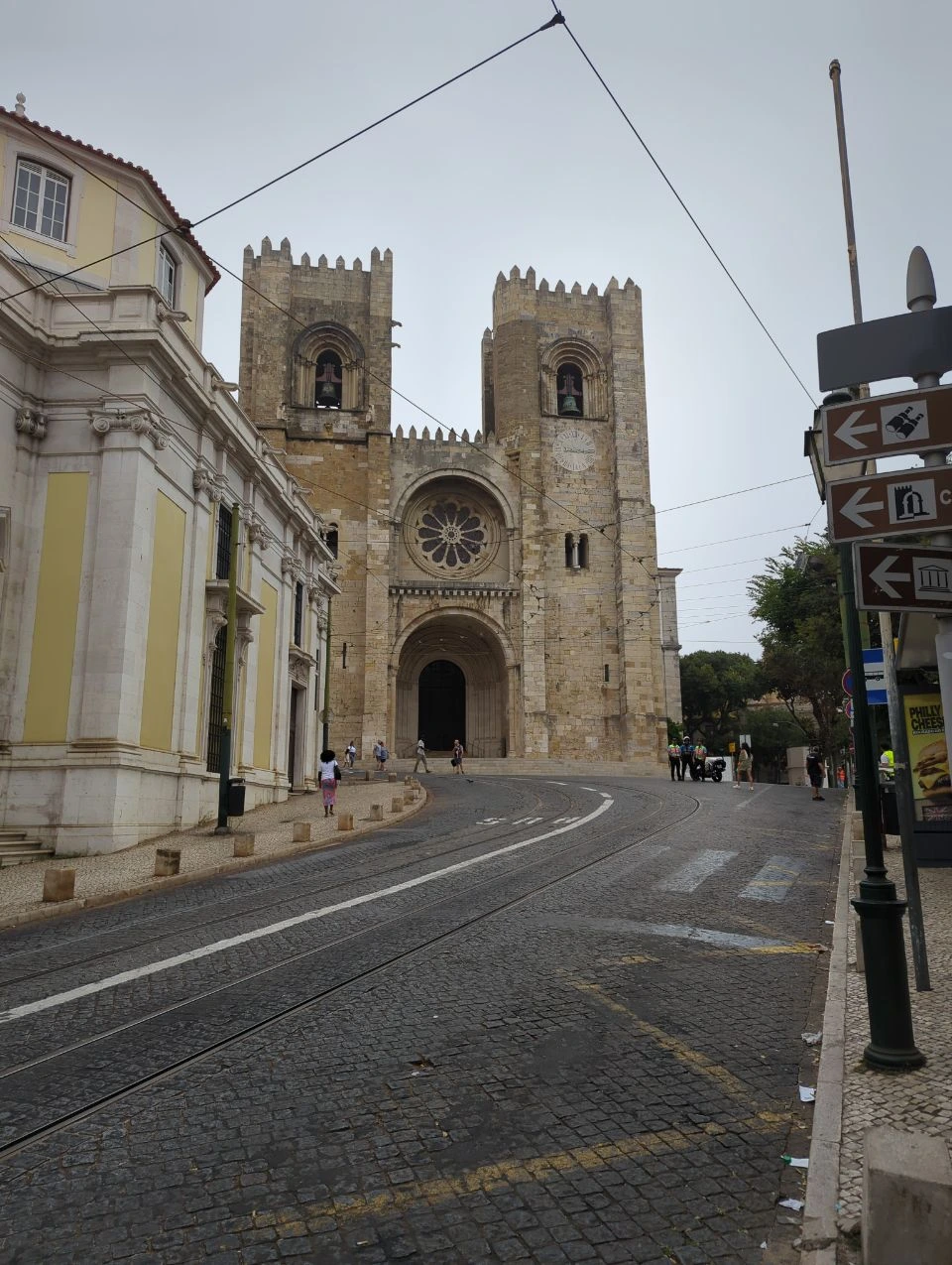 Lisbon Tram Photoshoot - Sé Cathedral Curve Sé Cathedral rises above a curved cobbled Lisbon street with tram rails, overhead wires and distant pedestrians.