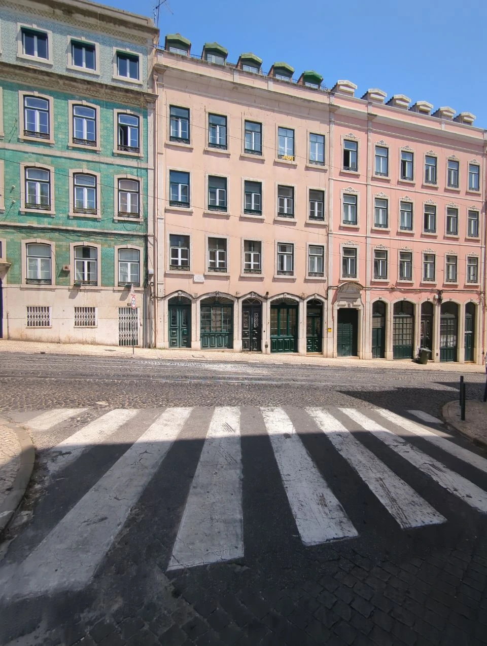 Green and pink façades face a zebra crossing beside tram rails on a sunlit Lisbon street.