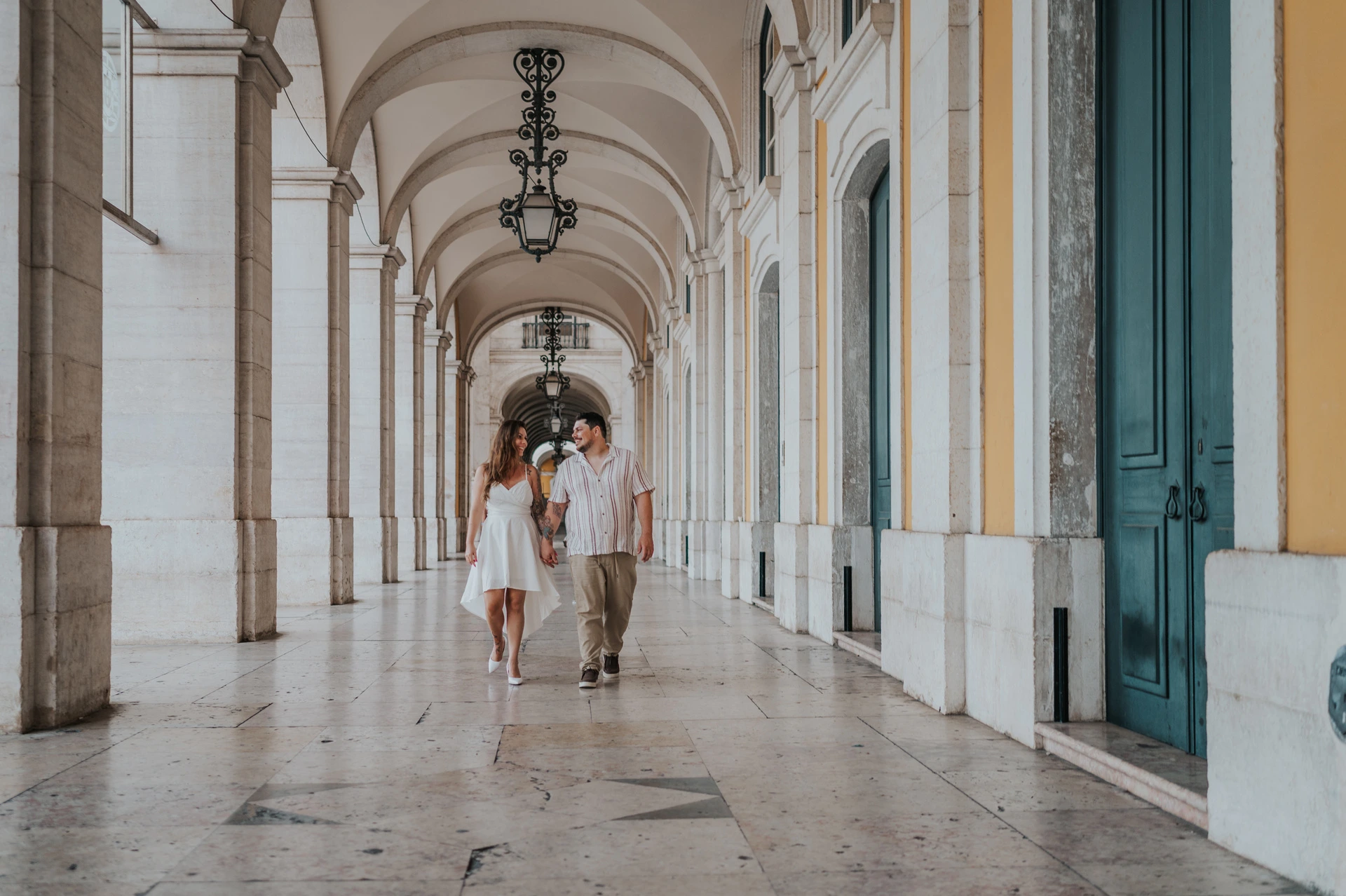 A wide view of the couple walking beneath the repeated arches and lanterns at Praça do Comércio during their couple photoshoot in Lisbon.