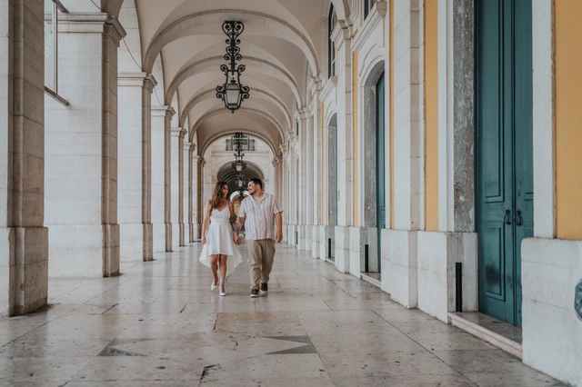 A wide view of the couple walking beneath the repeated arches and lanterns at Praça do Comércio during their couple photoshoot in Lisbon.