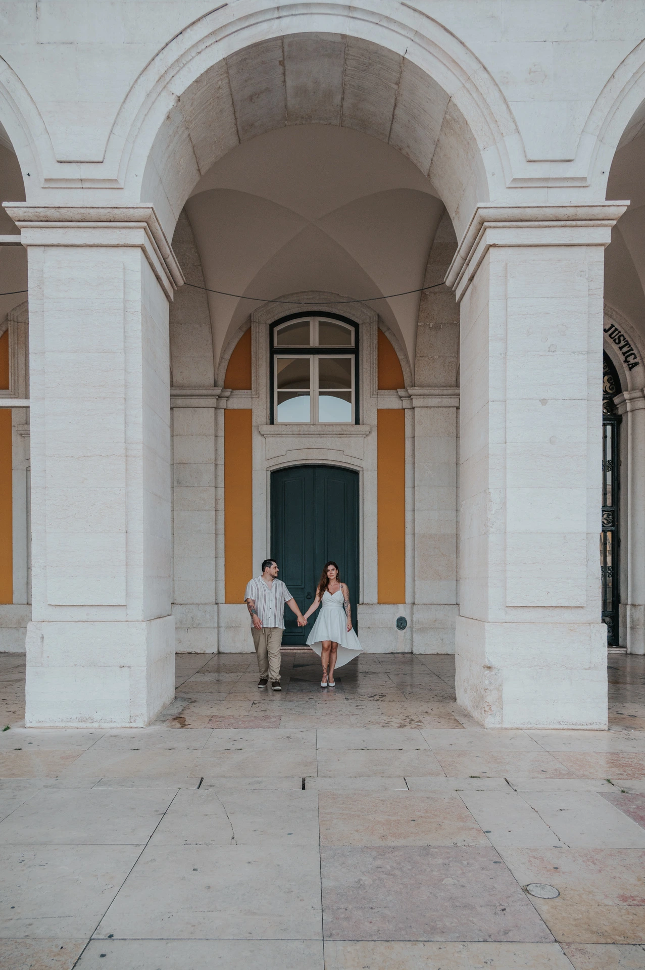 The couple standing hand in hand beneath a tall stone arch at Praça do Comércio during their couple photoshoot in Lisbon.