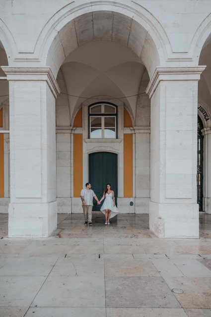 Hand in hand beneath a grand arch | Lisbon couple photoshoot The couple standing hand in hand beneath a tall stone arch at Praça do Comércio during their couple photoshoot in Lisbon.