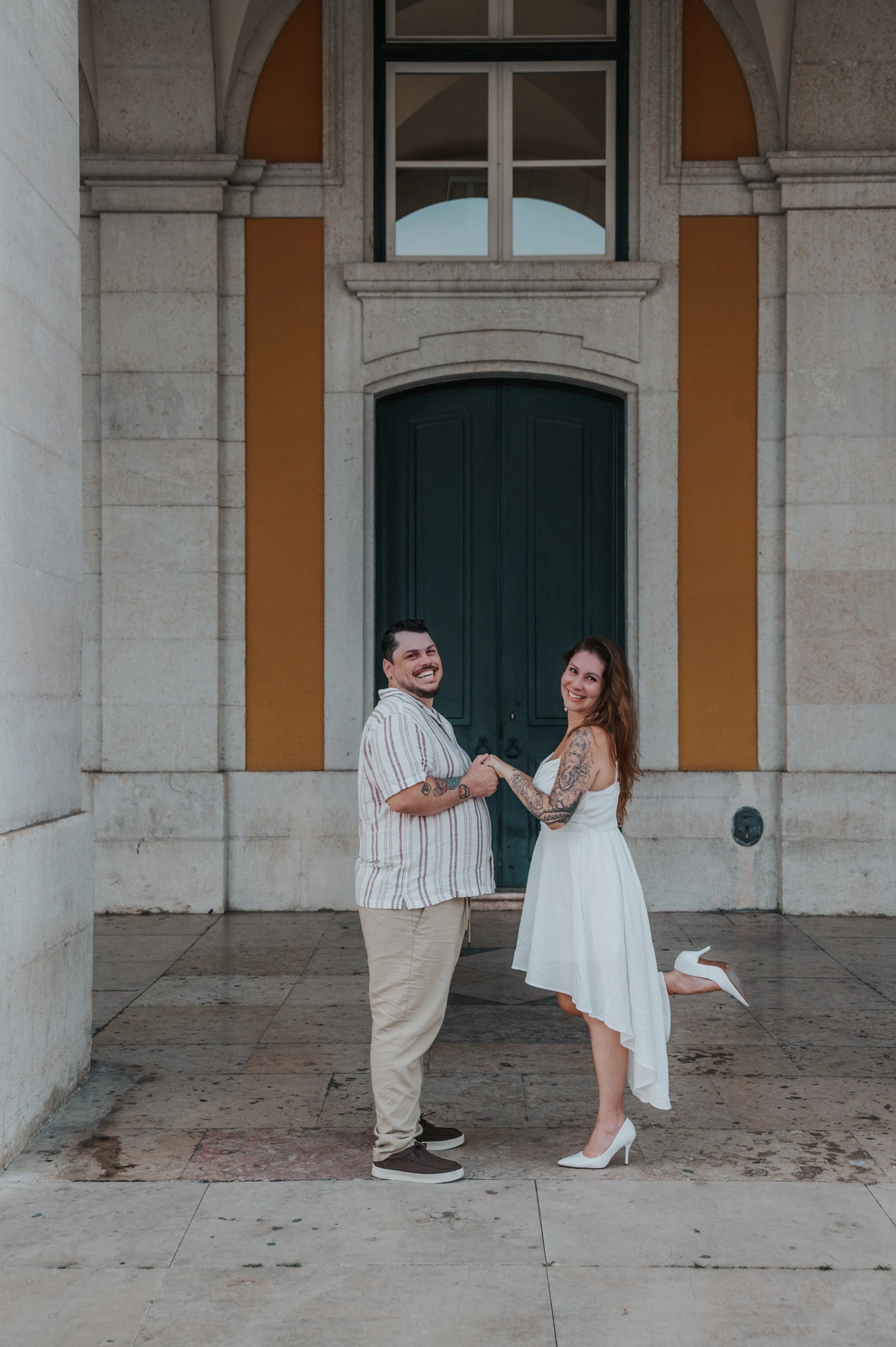 The couple holding hands as she turns towards the camera beneath the arcade during their couple photoshoot in Lisbon.