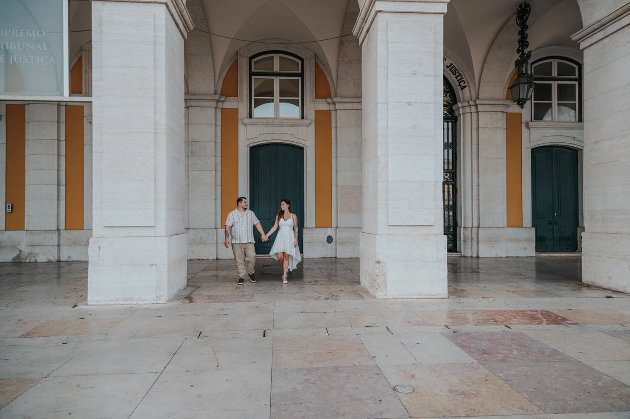 A wide portrait beneath the stone arches | Lisbon couple photoshoot A wide view of the couple holding hands between the large stone columns at Praça do Comércio during their couple photoshoot in Lisbon.