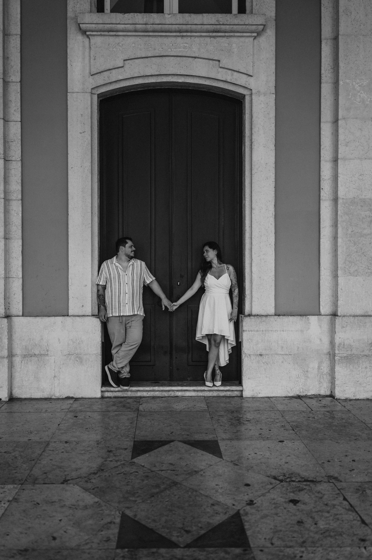 A black-and-white portrait of the couple holding hands across the doorway during their couple photoshoot in Lisbon.