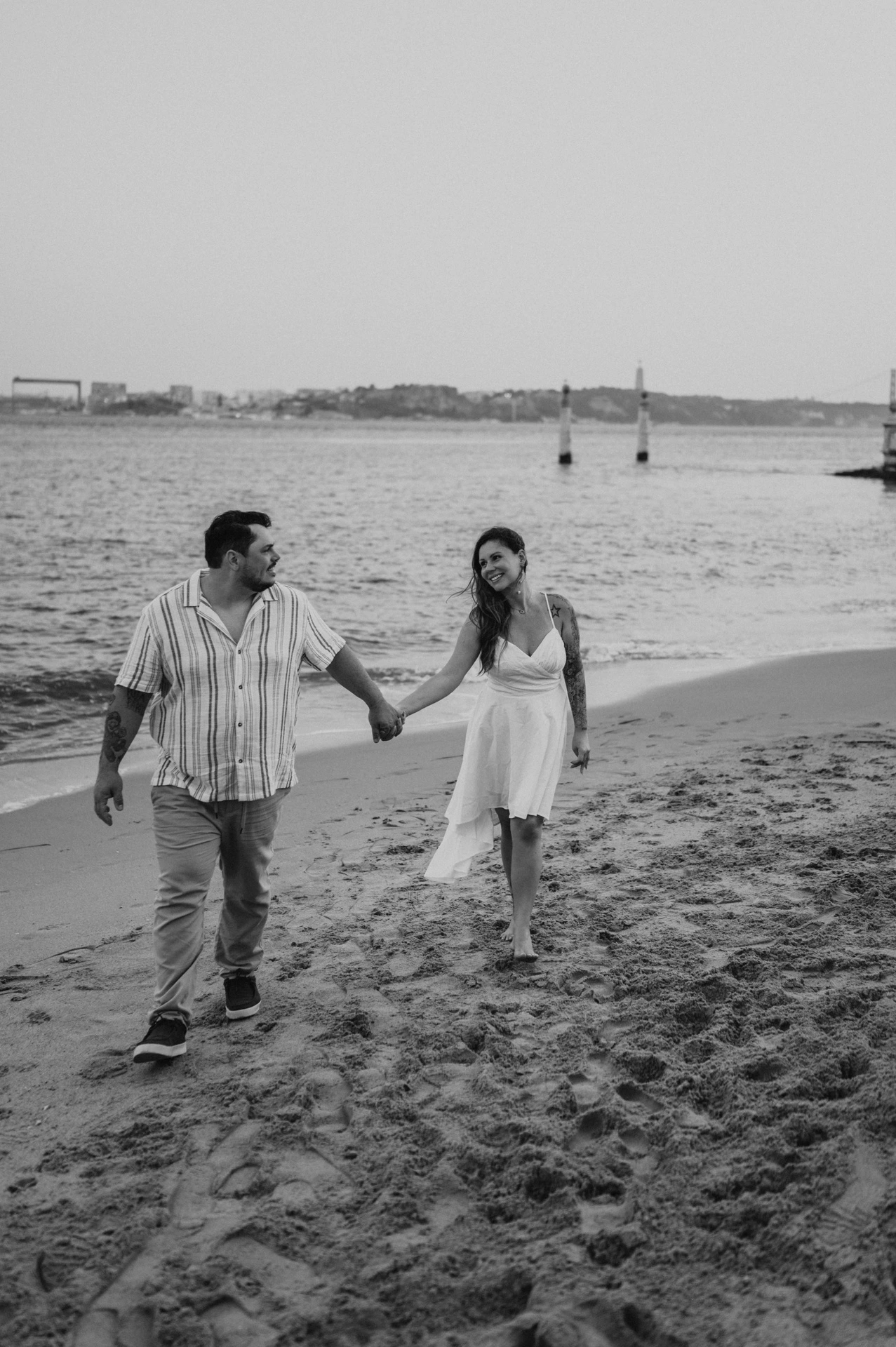 A black-and-white photograph of the couple walking hand in hand on the river beach during their couple photoshoot in Lisbon.