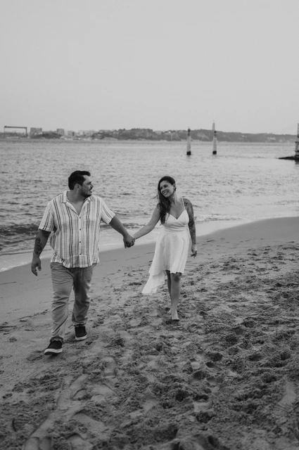 A river beach walk in black and white | Lisbon couple photoshoot A black-and-white photograph of the couple walking hand in hand on the river beach during their couple photoshoot in Lisbon.