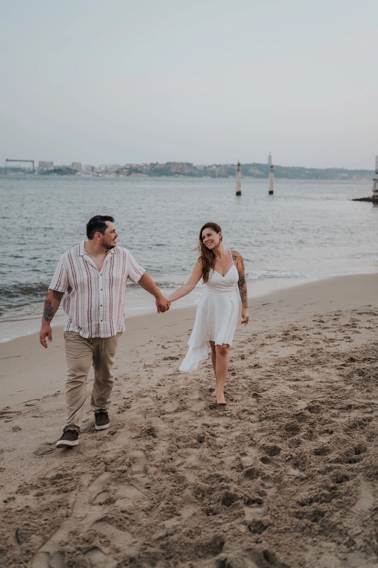 The couple smiling at each other as they walk along the sandy riverbank during their couple photoshoot in Lisbon.