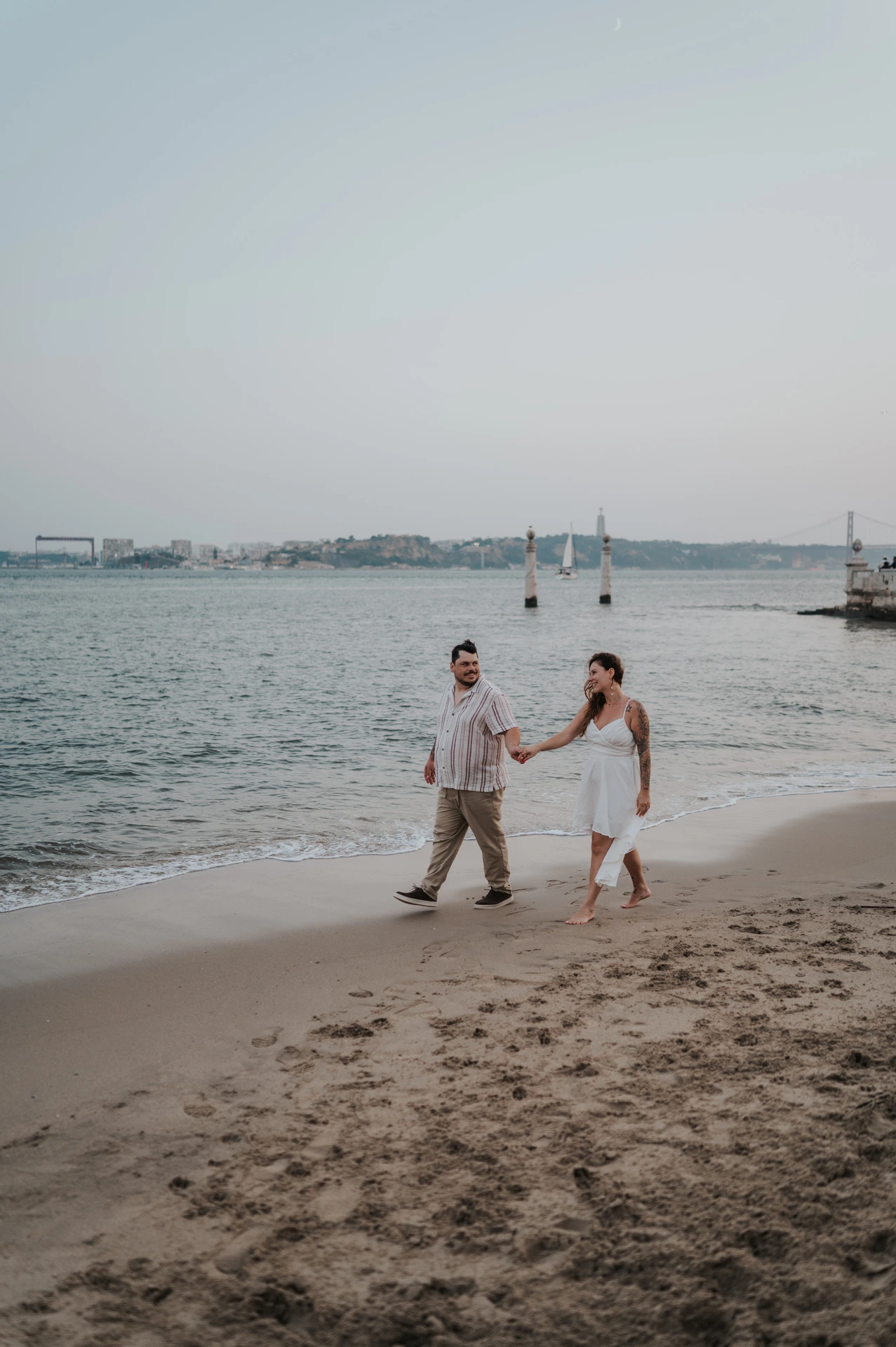 A distant portrait of the couple holding hands on the sand beside the river during their couple photoshoot in Lisbon.