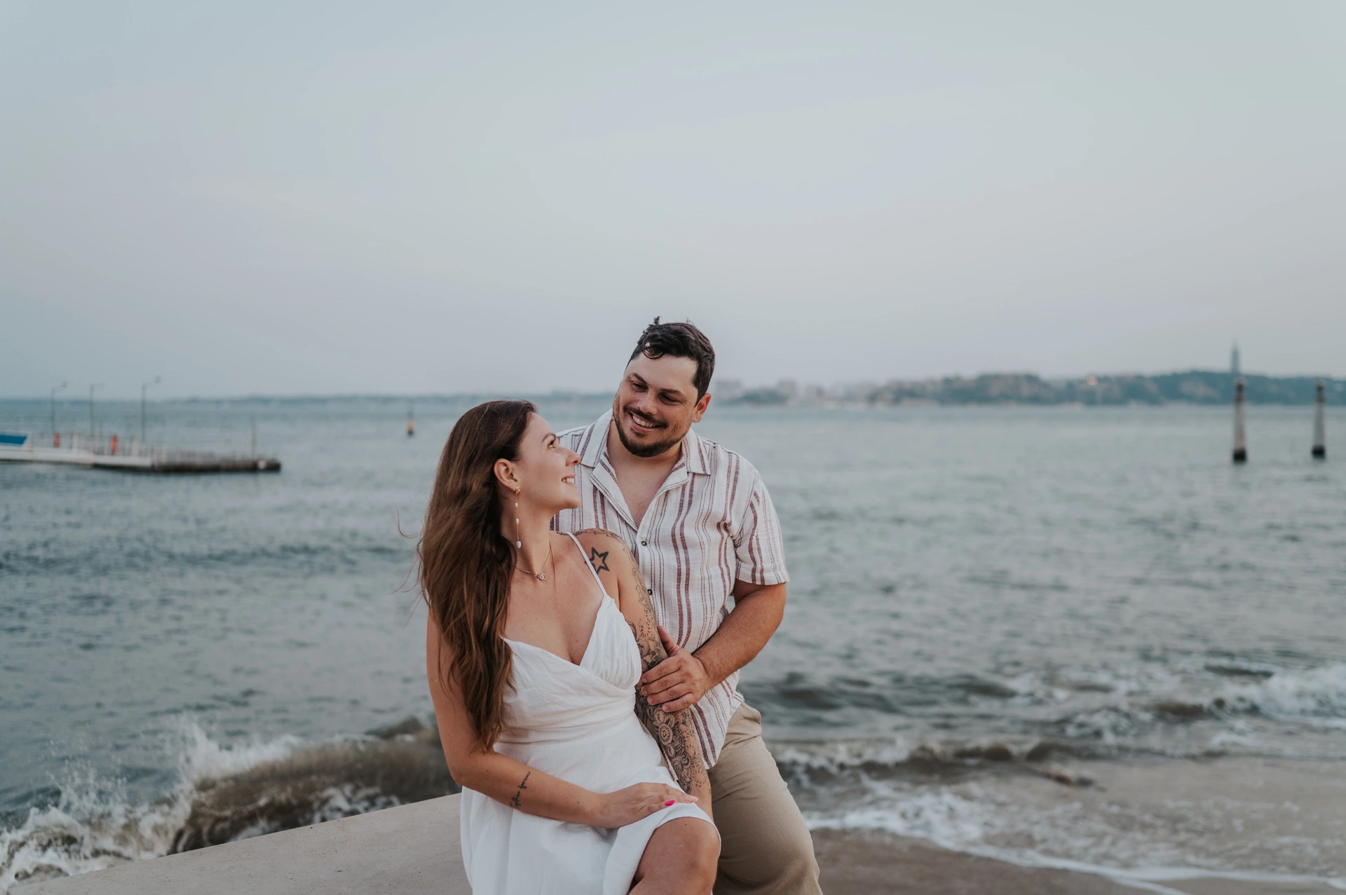 A shared smile beside the water | Lisbon couple photoshoot The couple smiling at one another beside the low waterfront wall and the water during their couple photoshoot in Lisbon.