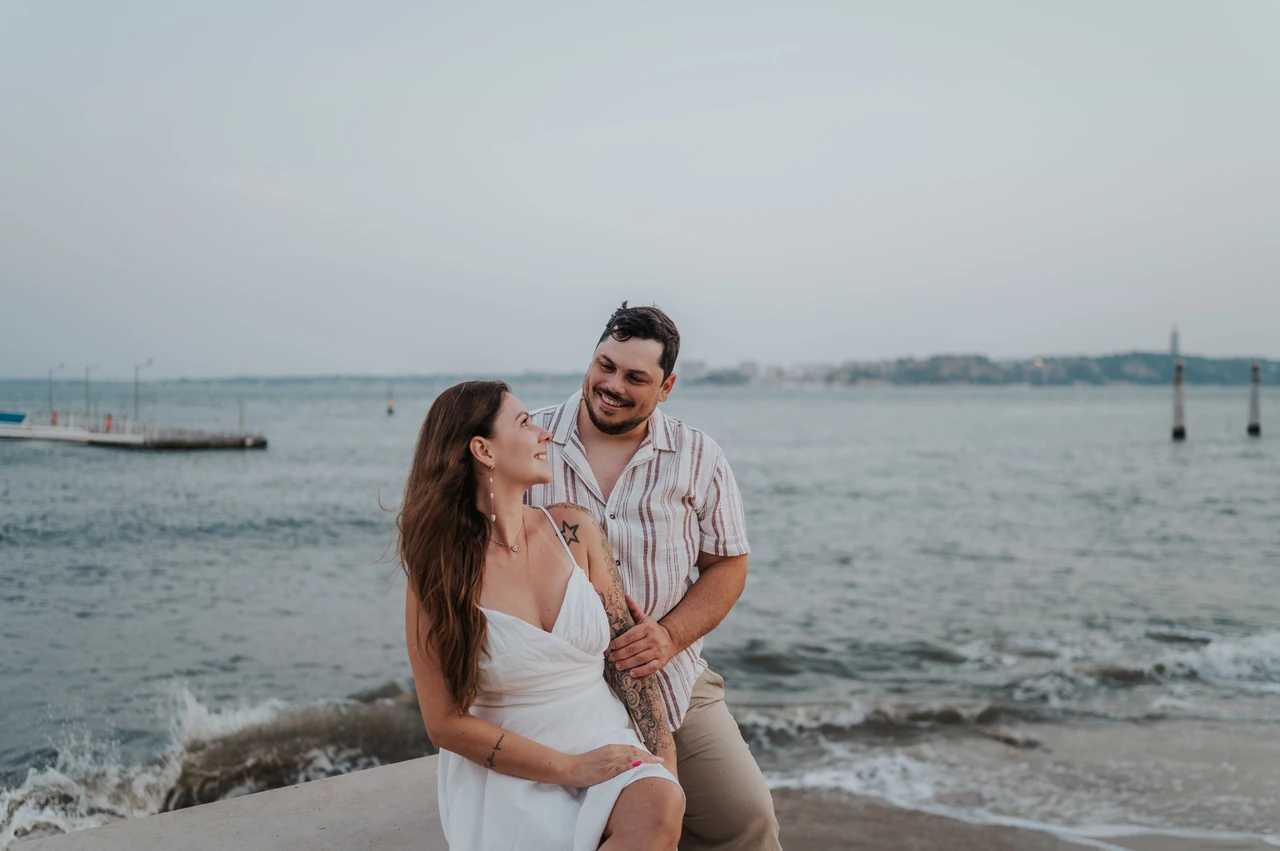A shared smile beside the water | Lisbon couple photoshoot The couple smiling at one another beside the low waterfront wall and the water during their couple photoshoot in Lisbon.