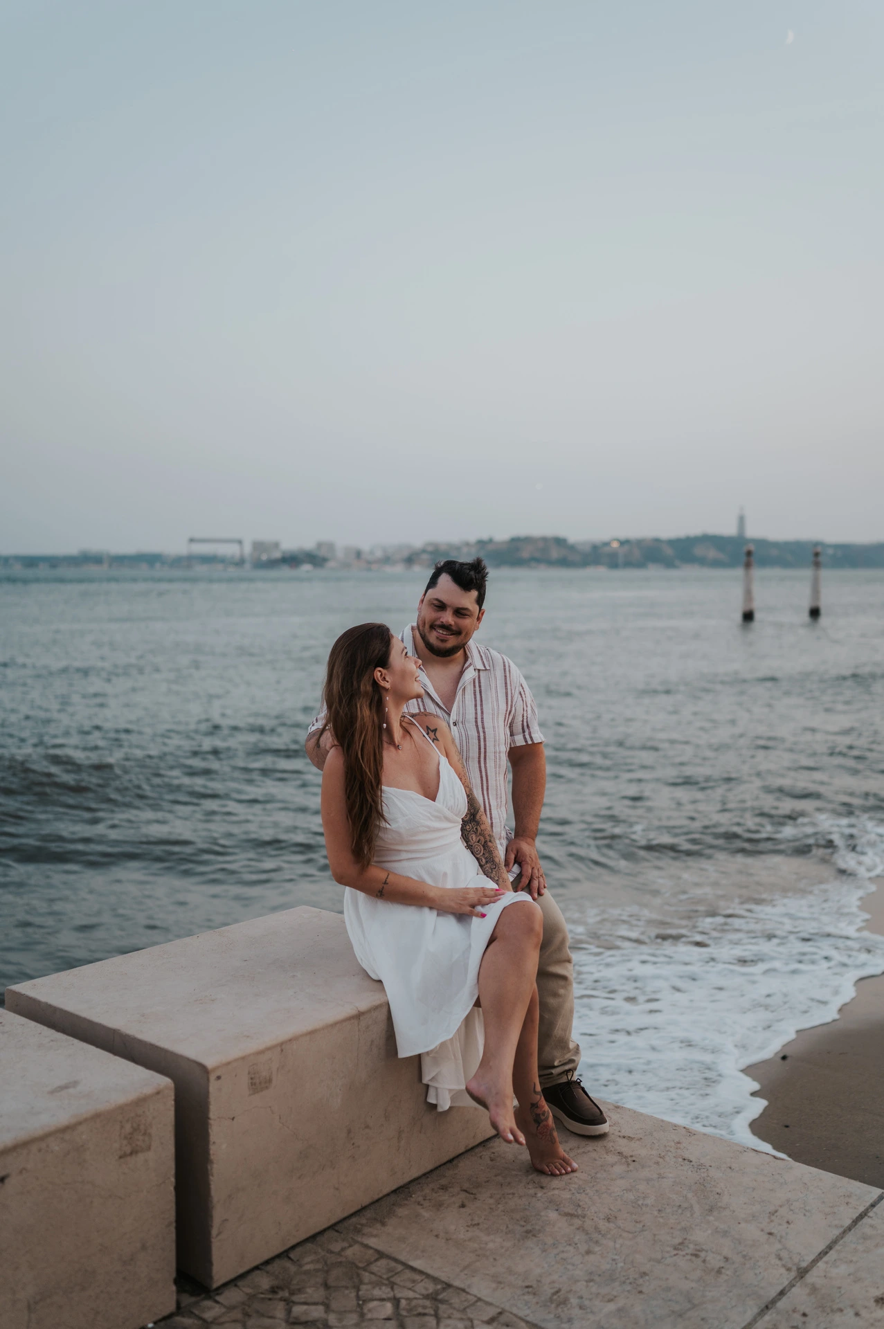 The woman seated on a low waterfront wall with her partner standing close behind her during their couple photoshoot in Lisbon.