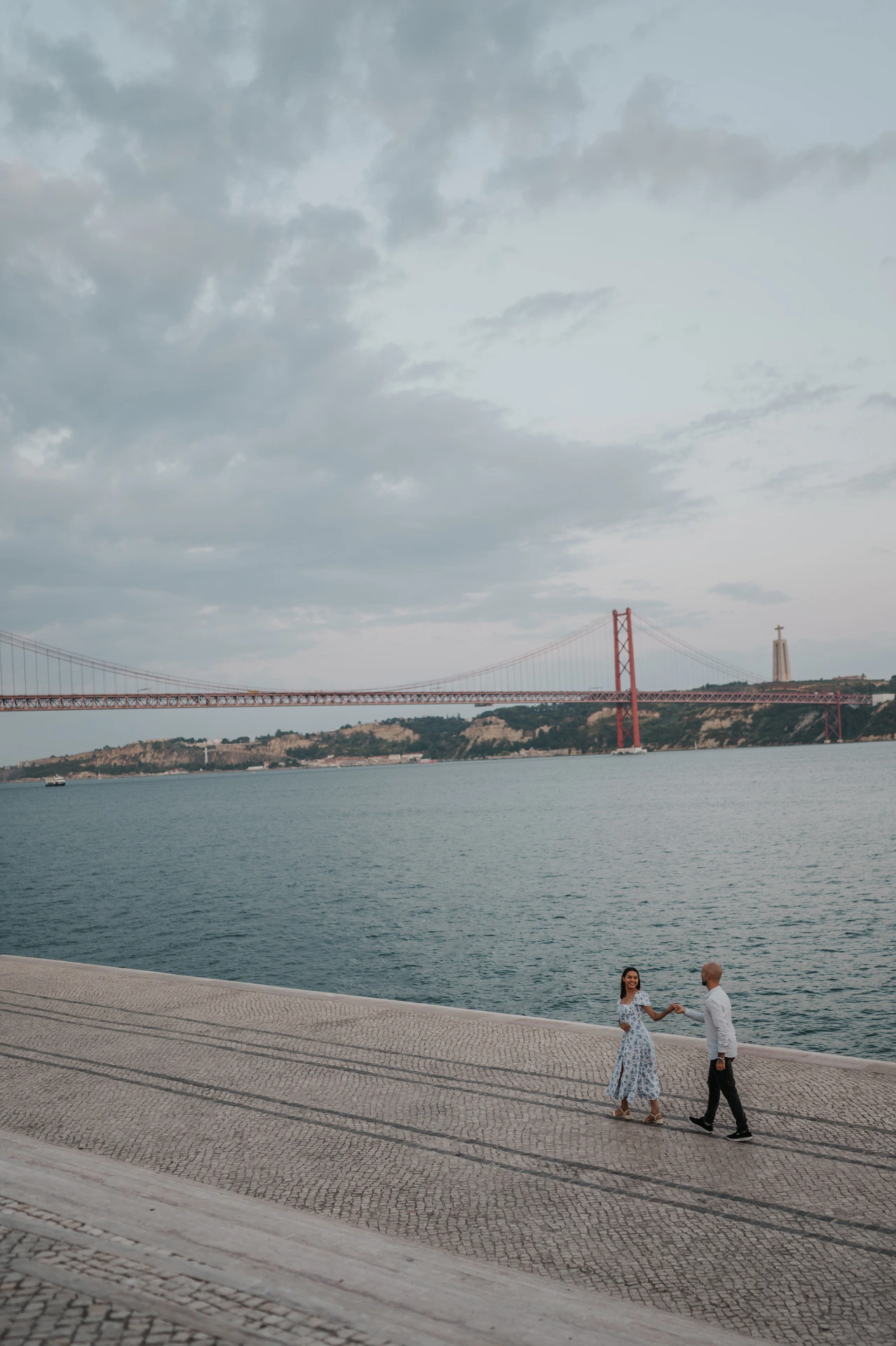 The couple holding hands beside the river with open water and clouds filling the view during their couple photoshoot in Lisbon.