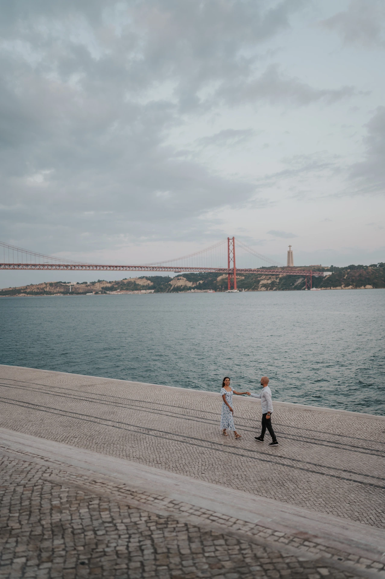An elevated view of the couple holding hands beside the river beneath an evening sky during their couple photoshoot in Lisbon.